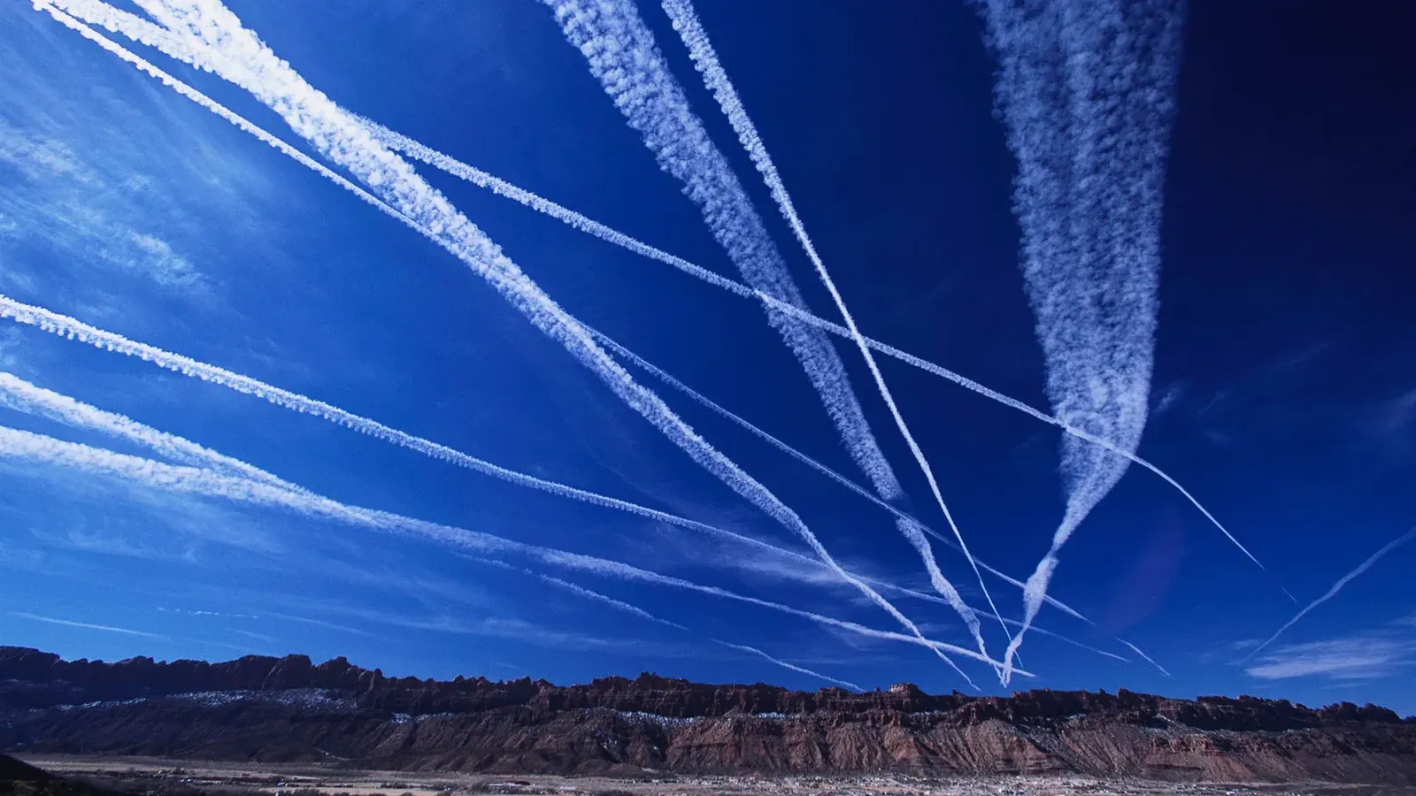 Multiple white contrails crossing a deep blue sky above a desert ridge.