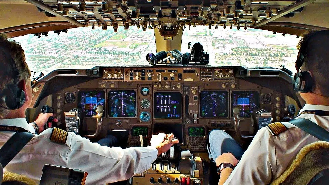 The flight deck of an airliner on approach, two pilots at the controls with a city below.